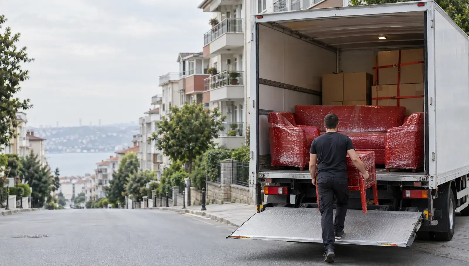 Sabah saatinde İstanbul sokağında açık kasalı beyaz kamyon; kasada kırmızı koruyucu ambalajla sarılmış koltuklar ve koliler, rampada sırtı dönük bir taşıyıcı